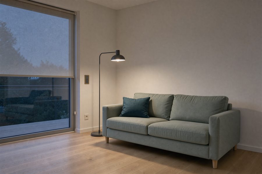 Wohnzimmer am Abend: graugrünes Sofa, eine eingeschaltete Stehlampe, halb heruntergelassenes Leinenrollo vor der Fensterfront, an der Wand ein Taster mit Messingrahmen.